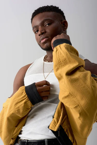 Low angle view of trendy afroamerican man in bomber jacket and tank top looking at camera while standing isolated on grey, contemporary shoot featuring stylish attire — Stock Photo