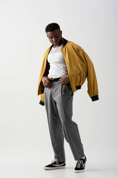 Selbstbewusster junger afrikanisch-amerikanischer Mann in Bomberjacke, der den Taillengürtel an der Hose berührt und auf grauem Hintergrund steht, zeitgenössisches Shooting mit stilvoller Kleidung — Stockfoto