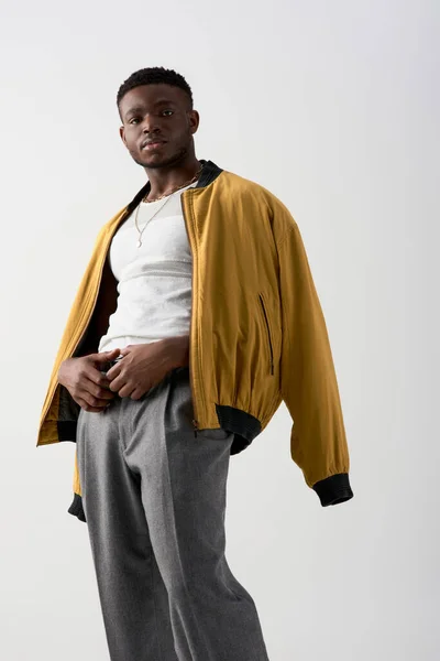 Впевнений молодий афроамериканець в костюмі бомбардувальника і штанах, який дивиться на камеру ізольовану на сірій, сучасній зйомці з стильним вбранням, модним твердженням. — Stock Photo