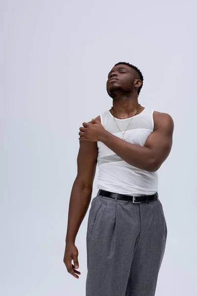 Vue en angle bas du jeune homme afro-américain détendu en t-shirt et pantalon sans manches touchant l'épaule isolé sur gris, pousse contemporaine avec une tenue élégante, musclé — Photo de stock