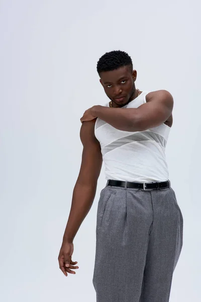 Selbstbewusster und muskulöser afrikanisch-amerikanischer Mann in ärmellosem T-Shirt und Hose, der die Schulter berührt und isoliert auf grauem, zeitgenössischem Shooting mit stylischer Kleidung in die Kamera blickt — Stockfoto