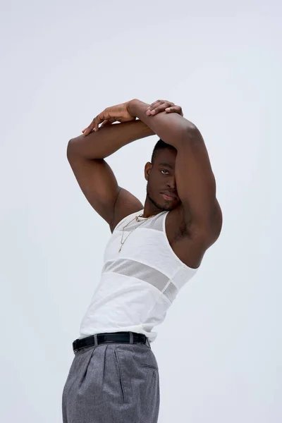 Homme musclé et jeune afro-américain en t-shirt et pantalon sans manches posant et regardant la caméra isolée sur gris, tournage contemporain avec une tenue élégante — Photo de stock