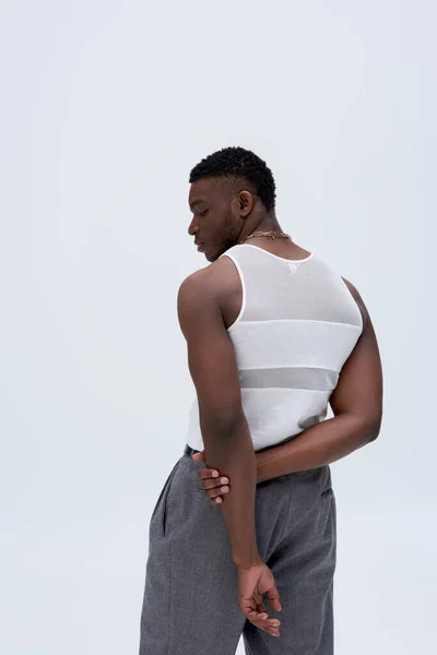 Vista lateral del joven afroamericano musculoso en pantalones y camiseta sin mangas posando con confianza en un atuendo elegante y de moda aislado en gris, bien parecido — Stock Photo