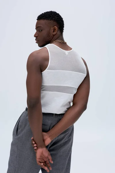 Vista lateral del atlético joven afroamericano en camiseta sin mangas y pantalones mirando hacia otro lado y posando con confianza en un atuendo elegante y de moda aislado en gris, bien parecido - foto de stock