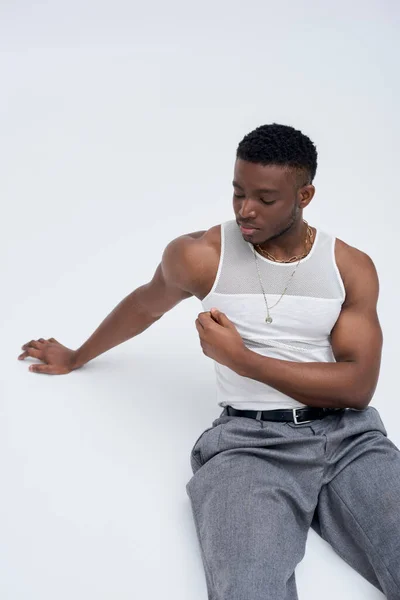 Fuerte modelo afroamericano en camiseta sin mangas y pantalones sentados y posando con confianza en un atuendo elegante y de moda sobre un fondo gris, atractivo, estilo callejero - foto de stock