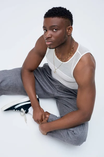 Retrato de hombre afroamericano musculoso y joven en pantalones y camiseta sin mangas sentado y posando con confianza en un atuendo elegante y de moda sobre fondo gris - foto de stock