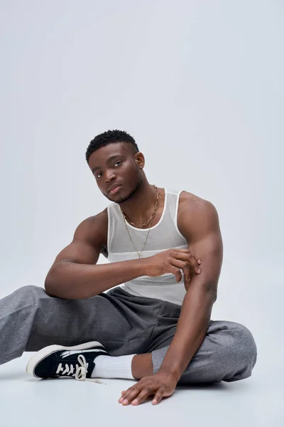 Young african american man in tank top and pants touching arm and posing confidently in stylish and trendy outfit on grey background, good looking, fashion sense — Stock Photo