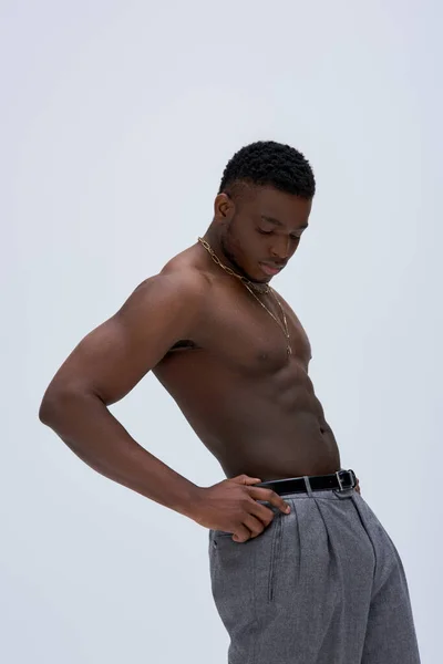 Homme afro-américain fort et torse nu en pantalon et colliers dorés debout isolé sur pose grise, confiante et moderne, pousse de mode, beau — Photo de stock