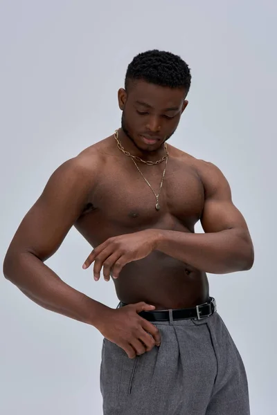 Good looking and shirtless afroamerican model in golden necklaces and pants looking at hand and standing isolated on grey, confident and modern pose, fashion shoot — Stock Photo