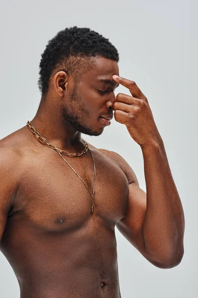 Shirtless and good looking african american model in golden necklaces touching nose isolated on grey, confident and modern pose, fashion shoot — Stock Photo
