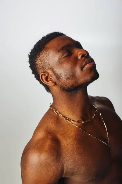 Homem americano africano relaxado e sem camisa com colares dourados fechando os olhos isolados em pose cinza, confiante e moderna, tiro de moda — Fotografia de Stock