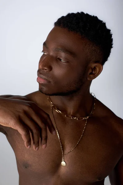 Retrato de hombre afroamericano joven y musculoso en collares dorados mirando hacia otro lado mientras está de pie aislado en pose gris, segura y moderna, sesión de fotos de moda - foto de stock