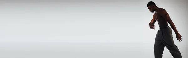 Vista lateral de modelo afroamericano muscular en pantalones y collares de pie en sombra aislada en pose gris, segura y moderna, sesión de moda, pancarta, hombre sin camisa - foto de stock