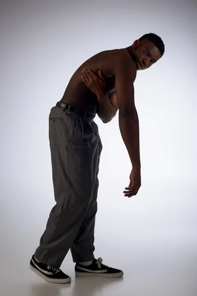 Pleine longueur de muscle et jeune modèle afro-américain en pantalon regardant la caméra sur fond gris, pose confiante et moderne, tournage de mode, homme torse nu — Photo de stock