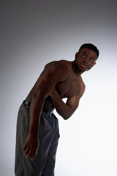 Безрубашечный и мускулистый африканский американец в ожерельях и брюках, смотрящий в сторону изолированный на серой, уверенной и современной позе, модная съемка, модель без рубашки — стоковое фото