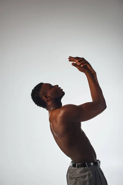 Вид сбоку молодого и мускулистого африканского мужчины в брюках, позирующего и стоящего в одиночестве на серой, уверенной и современной позе, модная фотосессия, безупречная модель — стоковое фото