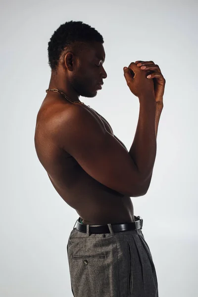 Vista lateral del hombre afroamericano relajado y sin camisa en collares y pantalones de pie aislados en pose gris, segura y moderna, sesión de moda, modelo muscular - foto de stock