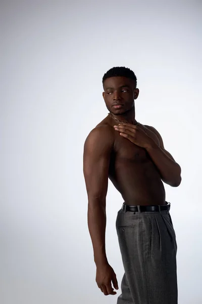 Homme afro-américain torse nu et jeune en colliers et pantalons dorés se tenant dans l'ombre isolé sur la pose grise, confiante et moderne, prise de vue mode, modèle musculaire — Photo de stock