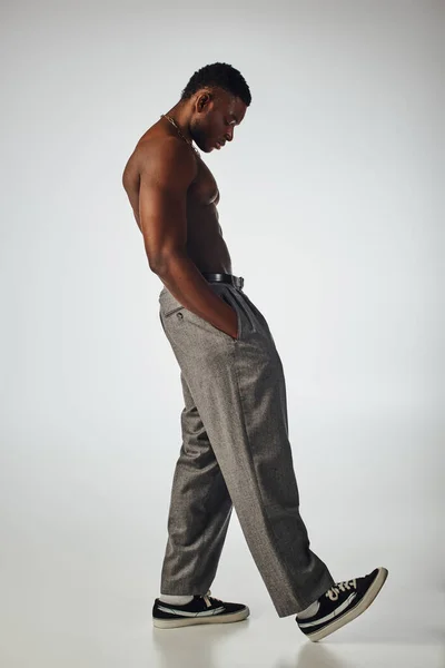 Side view of muscular afroamerican man in golden necklaces and pants holding hand in pocket on grey background, confident and modern pose, fashion shoot, shirtless model — Stock Photo
