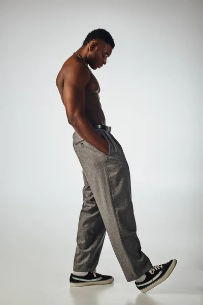 Vista lateral do homem afroamericano muscular em colares dourados e calças de mão no bolso em fundo cinza, pose confiante e moderna, tiro de moda, modelo sem camisa — Fotografia de Stock