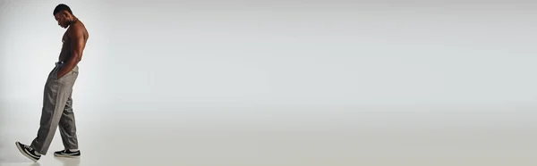 Longueur totale du modèle afroaméricain torse nu en colliers et pantalons dorés tenant la main dans la poche sur fond gris, pose confiante et moderne, prise de vue mode, bannière — Photo de stock