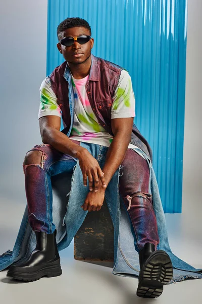 Homem americano africano na moda em óculos de sol e colete de ganga sentado em pedra e posando em cinza com folha de policarbonato azul no fundo, tiro de moda, moda sustentável, roupas DIY — Fotografia de Stock