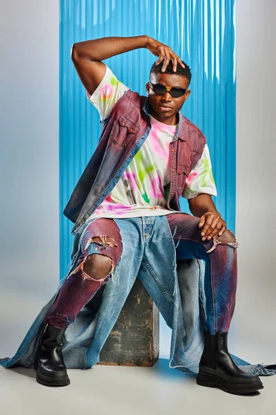 Stylish afroamerican man in sunglasses, denim vest and colorful t-shirt touching head while sitting on stone on grey with blue polycarbonate sheet at background, fashion shoot, sustainable fashion — Stock Photo