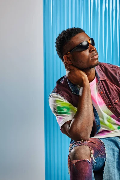 Homem afroamericano jovem na moda em óculos de sol, camiseta colorida e jeans rasgados tocando pescoço e posando em cinza com folha de policarbonato azul no fundo, tiro de moda, roupas DIY — Fotografia de Stock