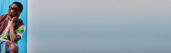 Jeune homme afro-américain à la mode en lunettes de soleil, gilet en denim coloré et jean déchiré touchant le cou sur fond gris avec une feuille de polycarbonate bleu, tournage de mode, bannière, vêtements DIY — Photo de stock