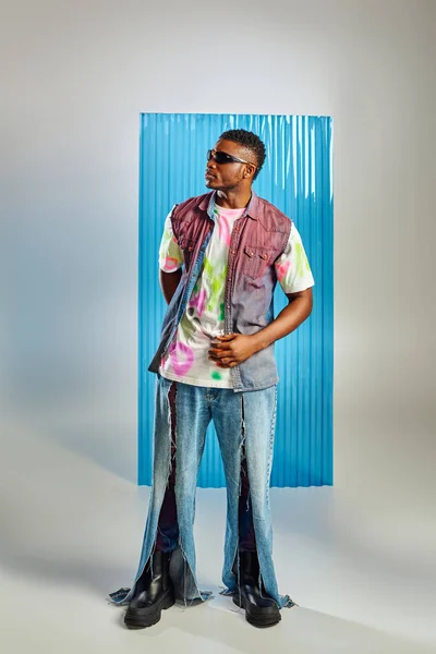 Comprimento total do moderno modelo afro-americano em camiseta colorida, óculos de sol e jeans rasgados em pé sobre cinza com folha de policarbonato azul no fundo, sessão de moda, estilo de vida sustentável — Fotografia de Stock