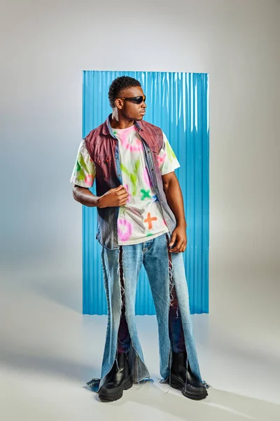 Comprimento total de homem afroamericano bonito e jovem em óculos de sol, colete de ganga e jeans rasgados em pé em cinza com folha de policarbonato azul no fundo, tiro de moda, roupas DIY — Fotografia de Stock