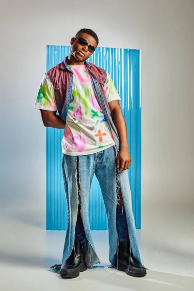Jeune homme afro-américain confiant en t-shirt coloré, lunettes de soleil et jeans déchiré debout sur gris avec une feuille de polycarbonate bleu à l'arrière-plan, tournage de mode, mode de vie durable — Photo de stock