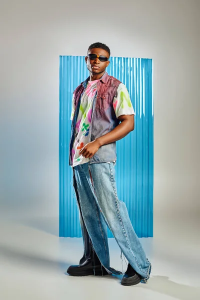 Повна довжина стильної молодої афроамериканської моделі в сонцезахисних окулярах, рвані джинси та джинсовий жилет, що стоїть на сірому з синім листом полікарбонату на фоні, модна стрілянина, одяг DIY — стокове фото