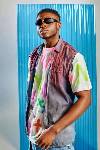 Portrait d'homme afro-américain à la mode en lunettes de soleil, t-shirt coloré et gilet en denim debout sur gris avec une feuille de polycarbonate bleu à l'arrière-plan, mode durable, vêtements DIY — Photo de stock