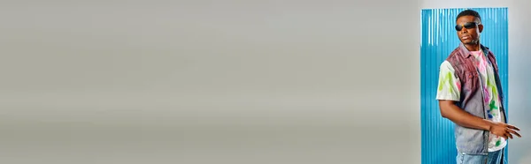 Jeune modèle afro-américain tendance en lunettes de soleil, t-shirt coloré et gilet en denim posant debout sur gris avec une feuille de polycarbonate bleu à l'arrière-plan, bannière, vêtements DIY — Photo de stock