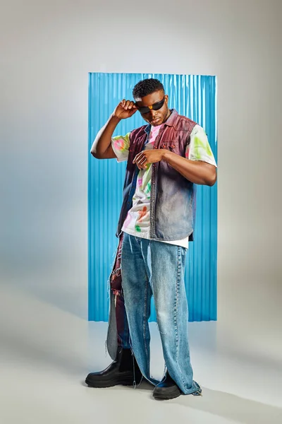 Longitud completa del modelo afroamericano joven de moda en gafas de sol, chaleco de mezclilla y camiseta colorida posando y de pie en gris con lámina de policarbonato azul en el fondo, moda sostenible - foto de stock