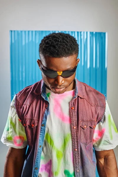 Retrato de hombre afroamericano joven y elegante con peinado de moda con gafas de sol, chaleco vaquero y camiseta colorida en gris con lámina de policarbonato azul en el fondo, moda sostenible - foto de stock
