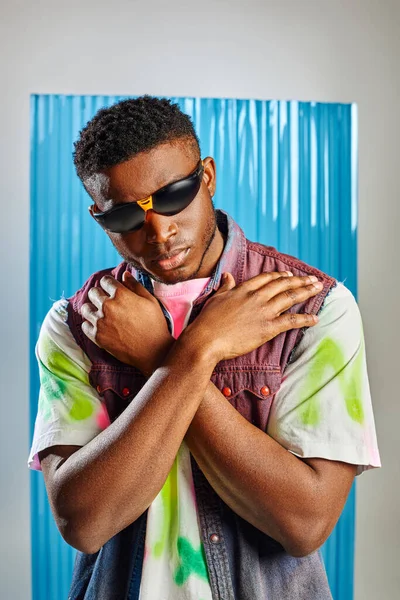 Elegante hombre afroamericano en gafas de sol, chaleco vaquero y colorida camiseta tocando hombros y de pie en gris con lámina de policarbonato azul en el fondo, moda sostenible, ropa de bricolaje - foto de stock