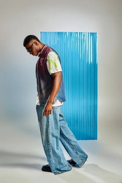 Vista lateral do modelo afroamericano na moda em óculos de sol, colete jeans e jeans andando em cinza com folha de policarbonato azul no fundo, moda sustentável, roupas DIY — Fotografia de Stock