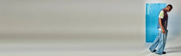 Vista laterale del giovane afroamericano alla moda in occhiali da sole, jeans e gilet in denim che cammina sul grigio con foglio di policarbonato blu sullo sfondo, servizio fotografico di moda, banner, moda sostenibile, abbigliamento fai da te — Foto stock