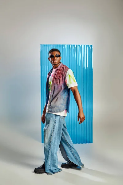Modelo afroamericano joven de moda en chaleco vaquero, camiseta colorida y jeans rasgados caminando sobre gris con sábana de policarbonato azul en el fondo, moda sostenible, ropa de bricolaje - foto de stock