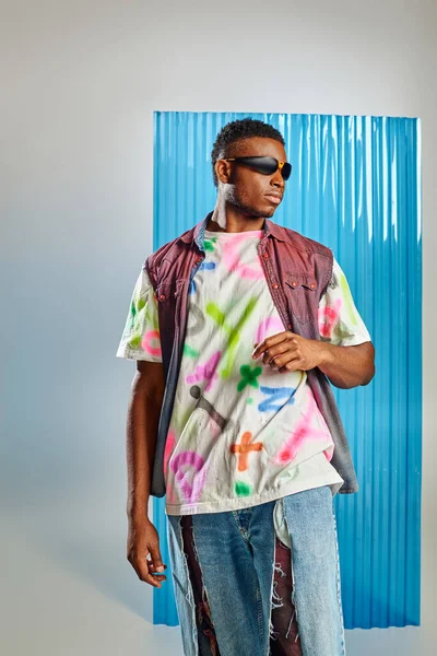 Модна молода афроамериканська модель в сонцезахисних окулярах, барвистий джинсовий жилет і рвані джинси, що стоять на сірому з синім полікарбонатним листом на фоні, стійка мода, одяг DIY — стокове фото