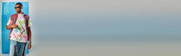 Jovem modelo afroamericano em colete de ganga colorido, óculos de sol e jeans rasgados elegantes em pé no cinza com folha de policarbonato azul no fundo, tiro de moda, banner, roupas DIY — Fotografia de Stock