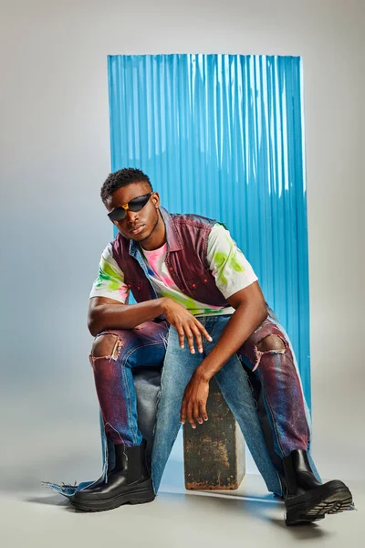 Modèle afro-américain tendance en lunettes de soleil, t-shirt coloré et jean déchiré assis sur fond gris avec une feuille de polycarbonate bleu, tournage de mode, mode durable, vêtements DIY — Photo de stock