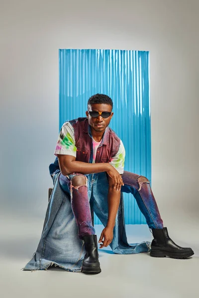 Modelo afroamericano em óculos de sol, colete de ganga colorido e jeans rasgado contemporâneo sentado em pedra em cinza com folha de policarbonato azul no fundo, moda sustentável, roupas DIY — Fotografia de Stock