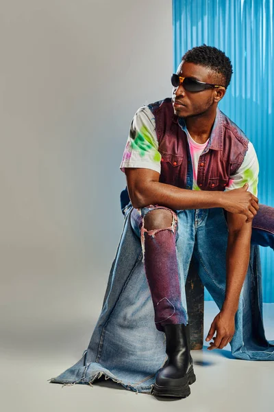 Hombre afroamericano confiado en gafas de sol, chaleco vaquero y vaqueros rasgados de moda sentado en piedra sobre gris con lámina de policarbonato azul en el fondo, moda sostenible, ropa de bricolaje - foto de stock