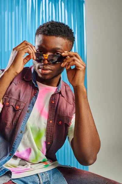 Portrait of stylish afroamerican man in colorful ripped jeans and denim vest holding sunglasses and looking at camera on grey with blue polycarbonate sheet at background, fashion shoot, DIY clothing — Stock Photo