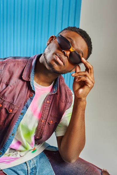 Modelo afroamericano jovem na moda em colete de ganga e camiseta colorida tocando óculos de sol em cinza com folha de policarbonato azul no fundo, tiro de moda, roupas DIY, estilo de vida sustentável — Fotografia de Stock