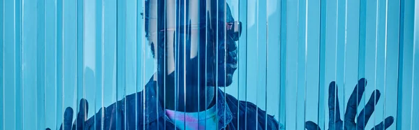 Modelo masculino afroamericano jovem na moda em óculos de sol e colete de ganga tocando folha de policarbonato azul, tiro de moda, bandeira, roupas DIY, estilo de vida sustentável — Fotografia de Stock