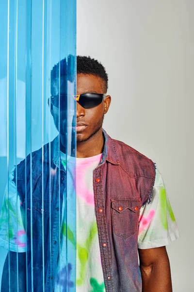 Portrait of fashionable young afroamerican man in sunglasses, denim vest and t-shirt standing behind blue polycarbonate sheet on grey background, fashion shoot, DIY clothing, sustainable lifestyle — Stock Photo